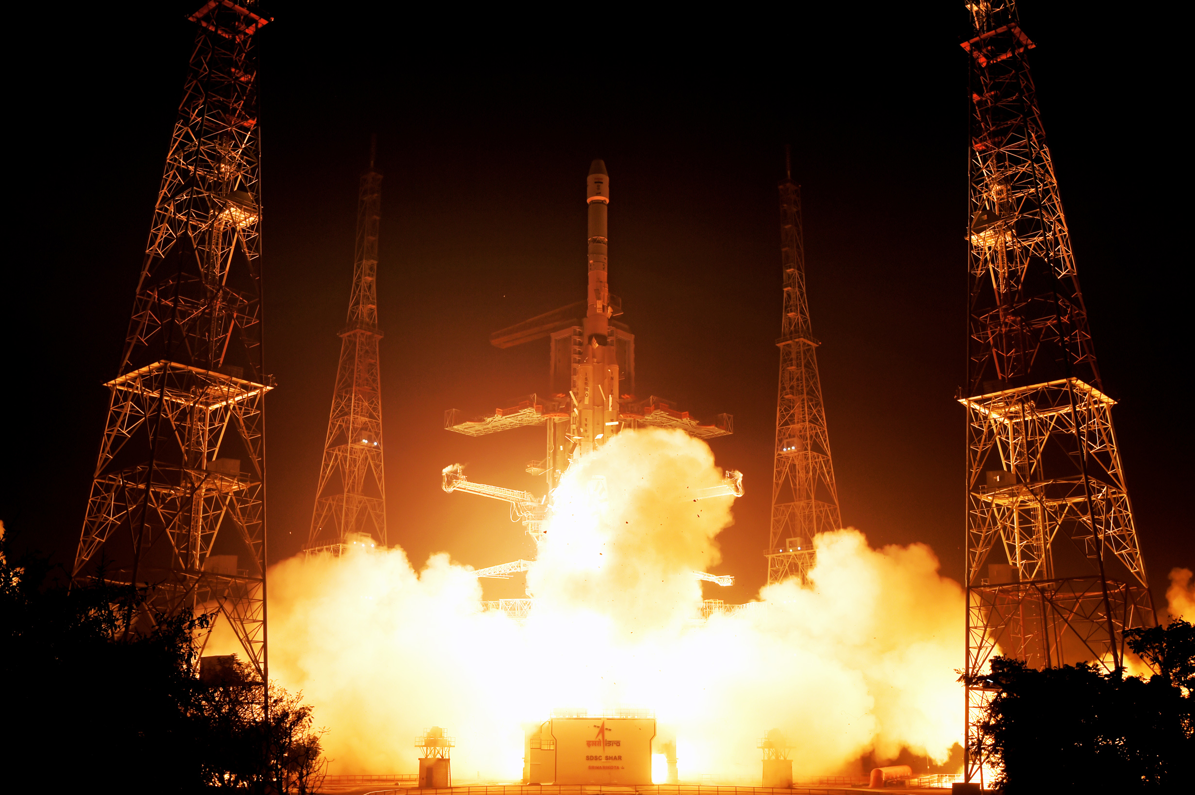 GSLV-F15 rises between illuminated launch towers at Sriharikota, with orange exhaust below the rocket.
